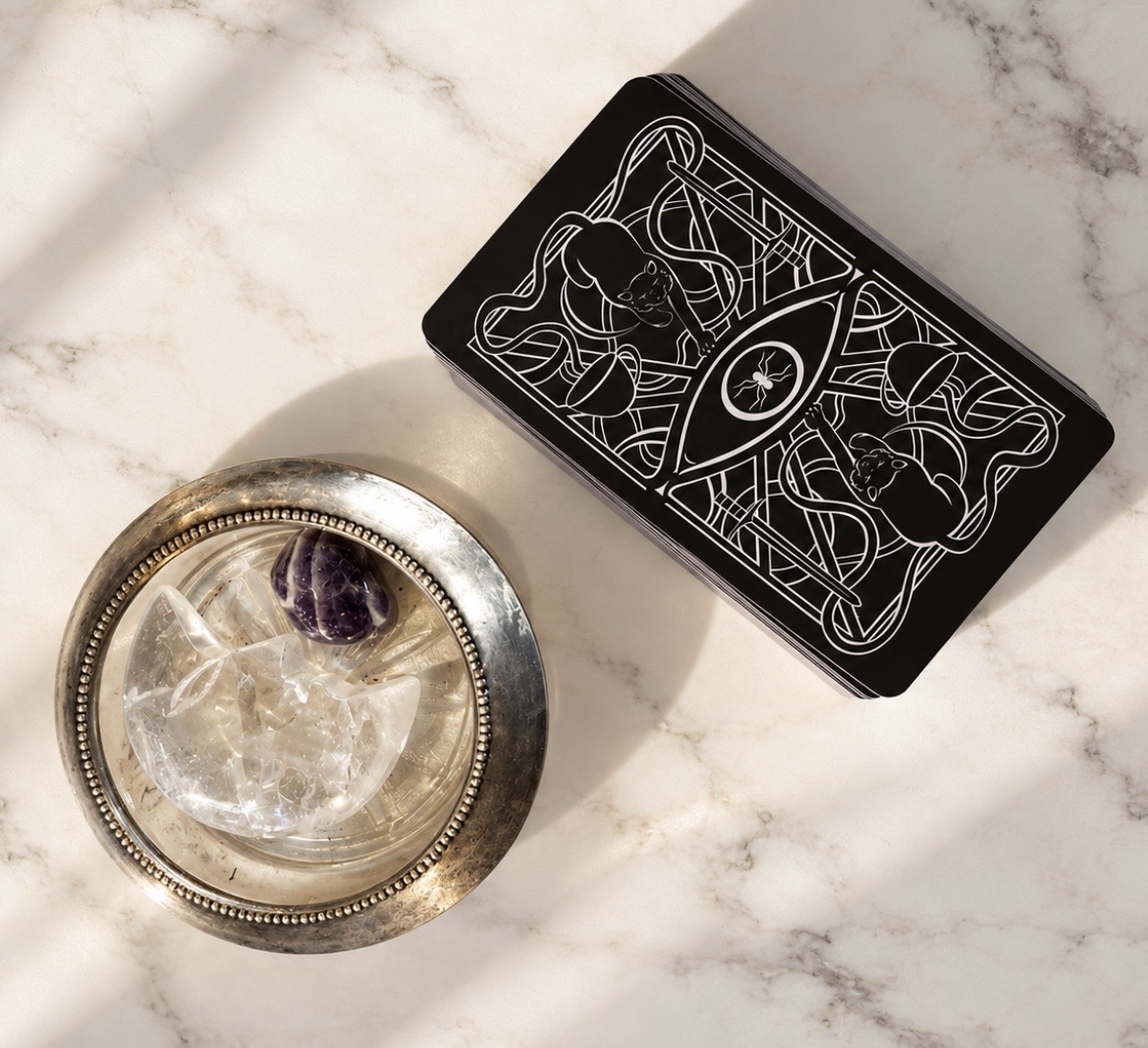 Black tarot deck and a silver dish of crystals on cream marble in soft sunlight