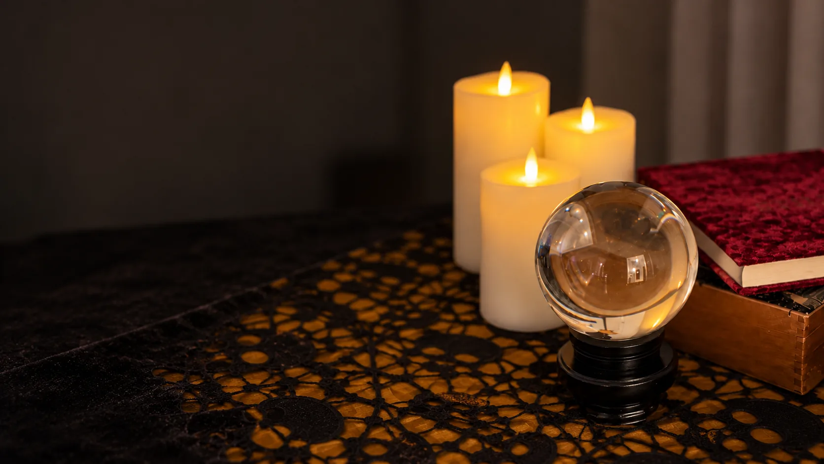 Clear crystal ball on a black stand beside pillar candles and a red velvet book on skull lace